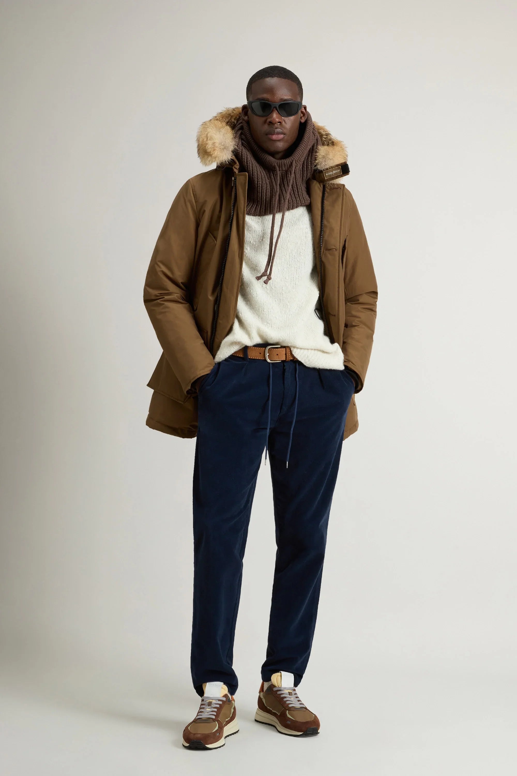 Modello uomo con giacca invernale marrone con cappuccio in pelliccia, maglione bianco, pantaloni blu e sneakers.