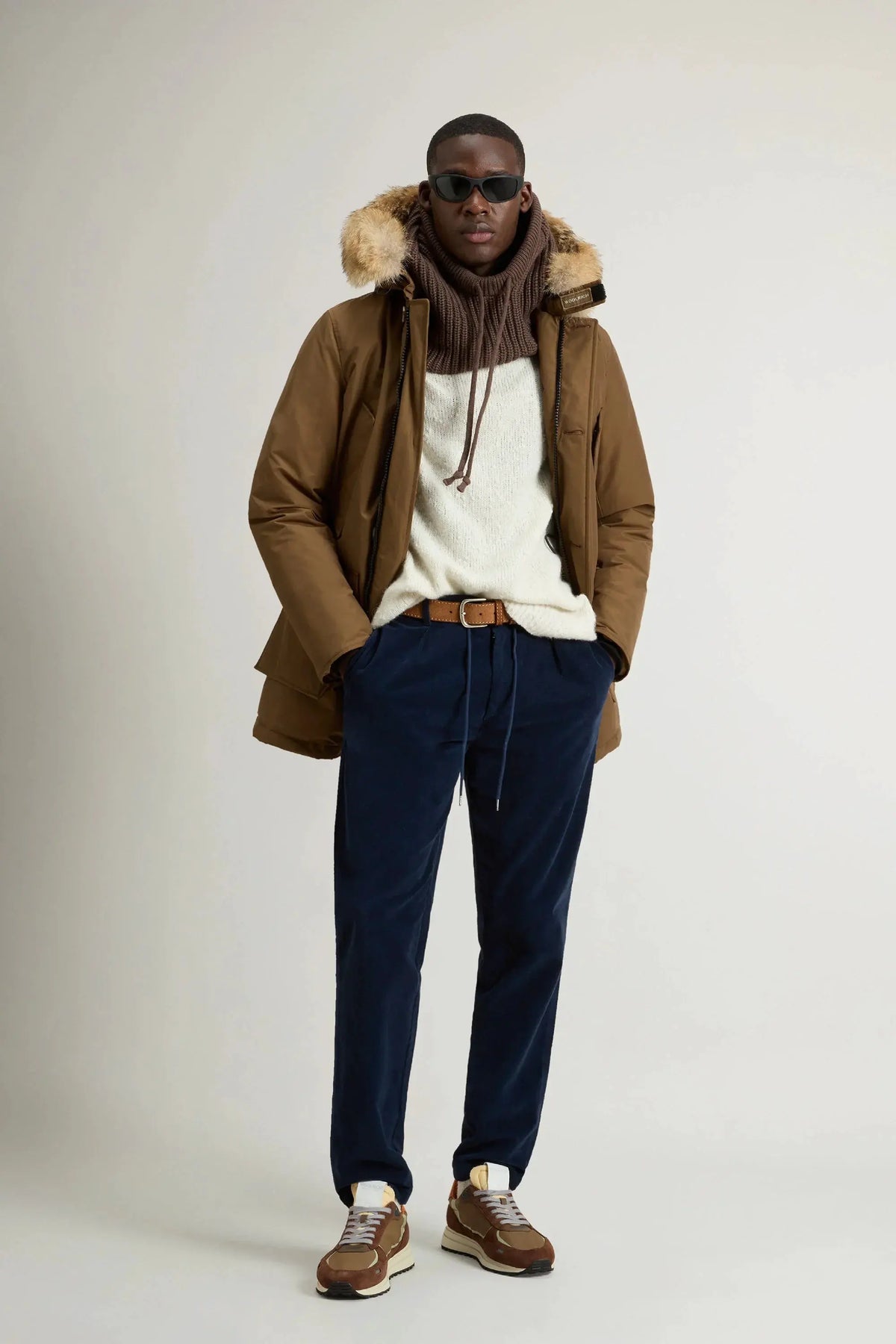 Modello uomo con giacca invernale marrone con cappuccio in pelliccia, maglione bianco, pantaloni blu e sneakers.