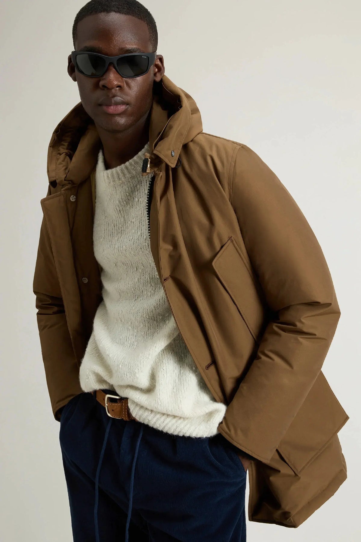 Uomo con giacca invernale marrone, maglione chiaro, pantaloni blu e occhiali da sole