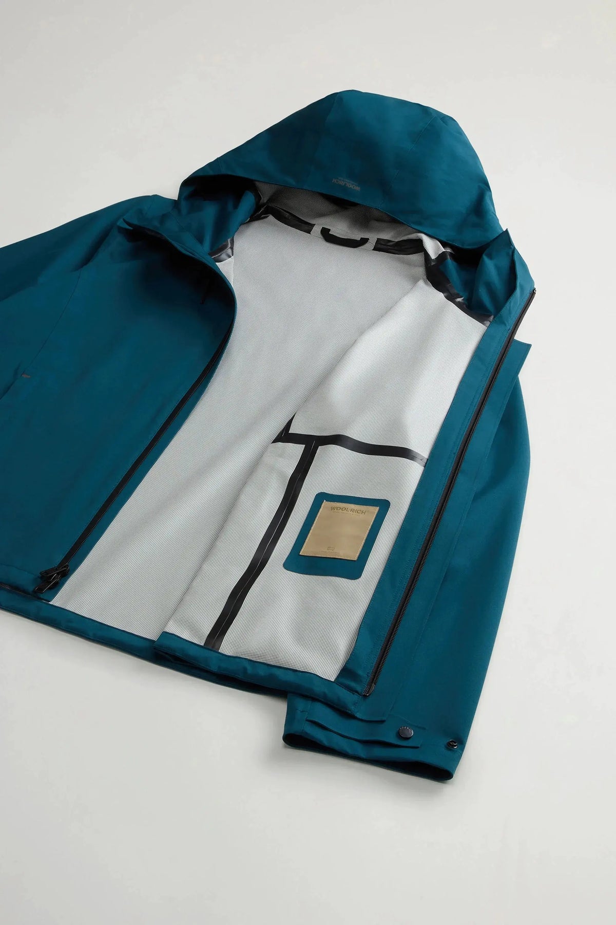 Giacca impermeabile blu petrolio con cappuccio, zip frontale e fodera interna bianca a vista.