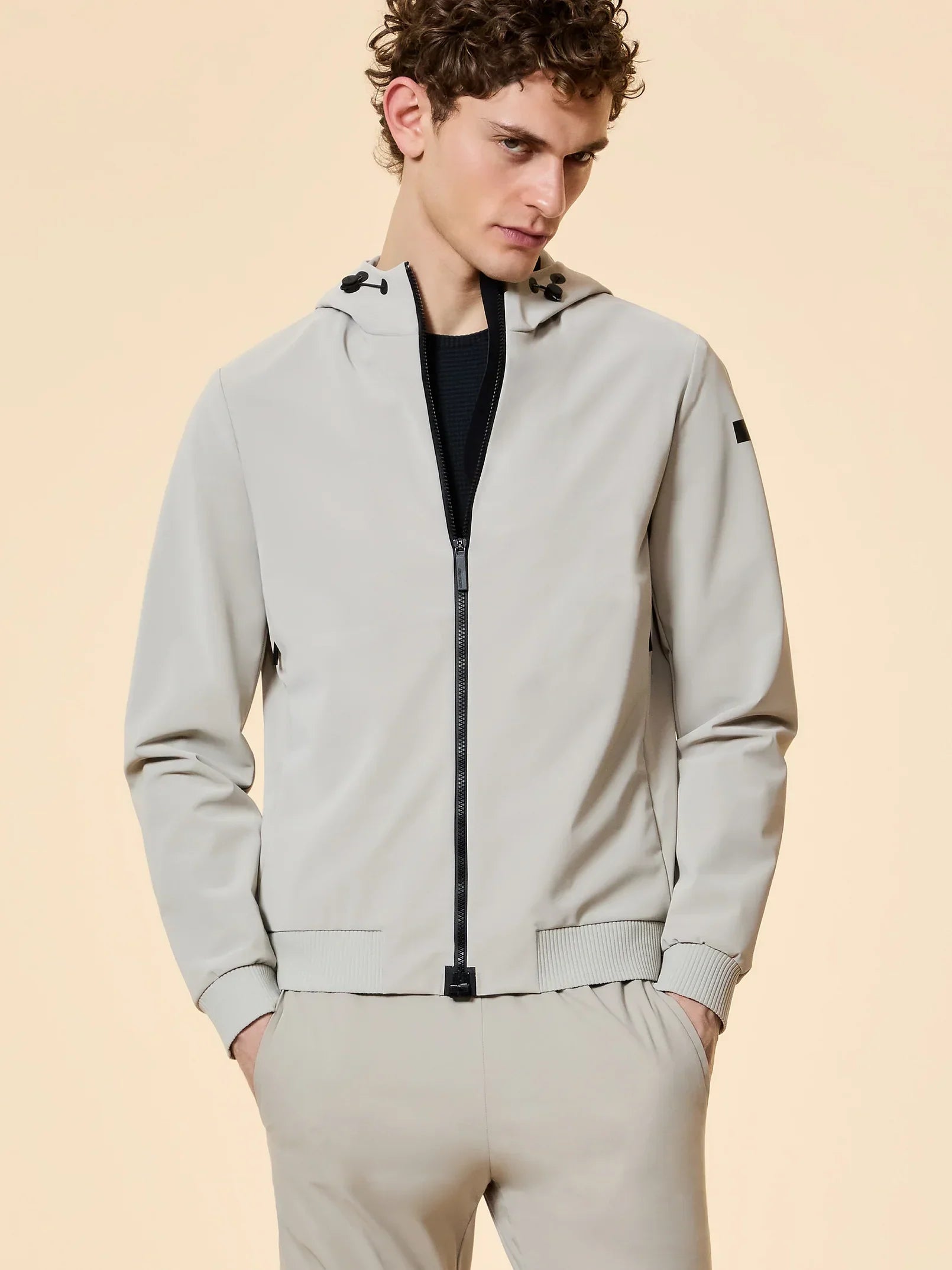 Giacca uomo beige con cappuccio, zip nera e pantaloni coordinati, stile casual elegante.