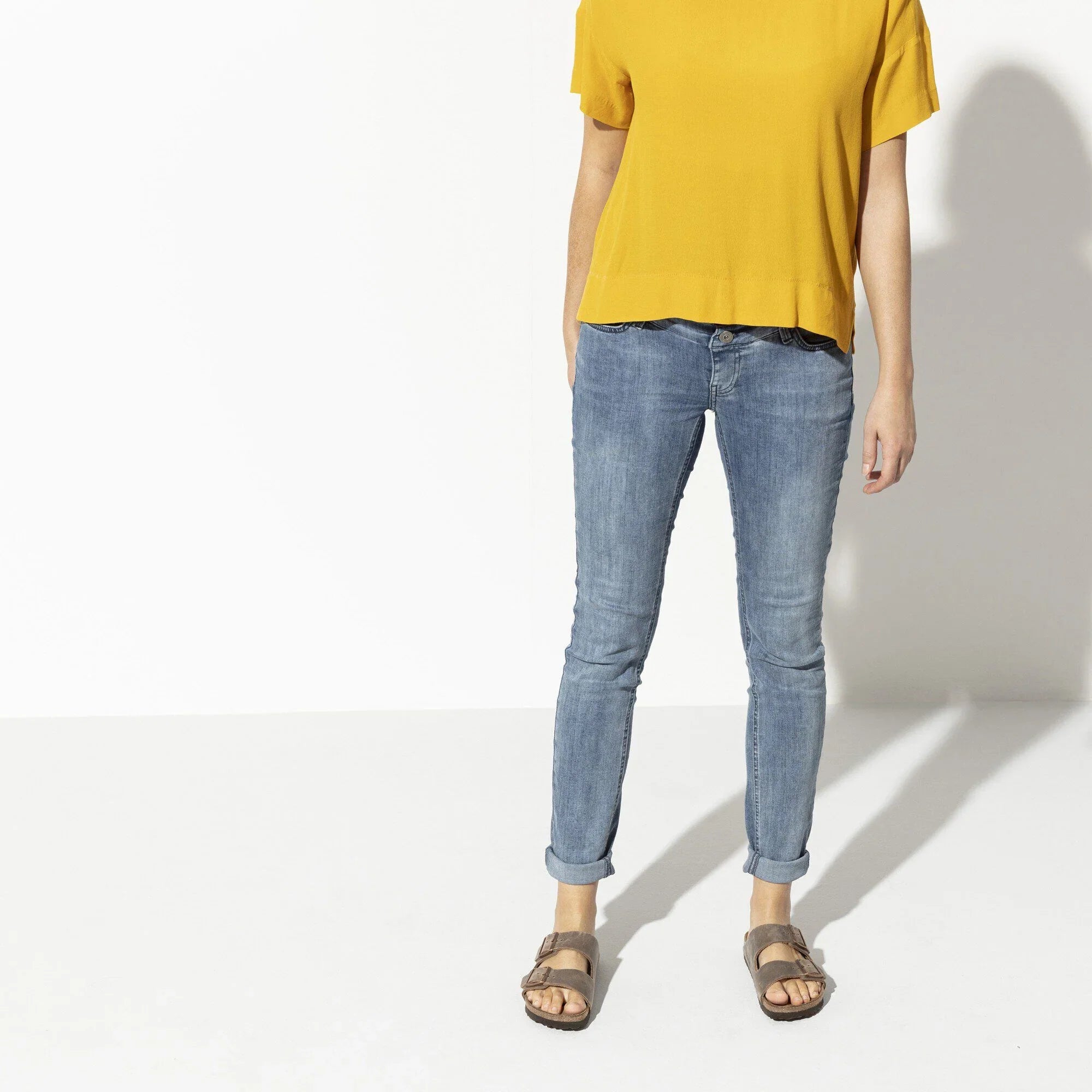 Donna con t-shirt gialla, jeans skinny e sandali marroni su sfondo chiaro
