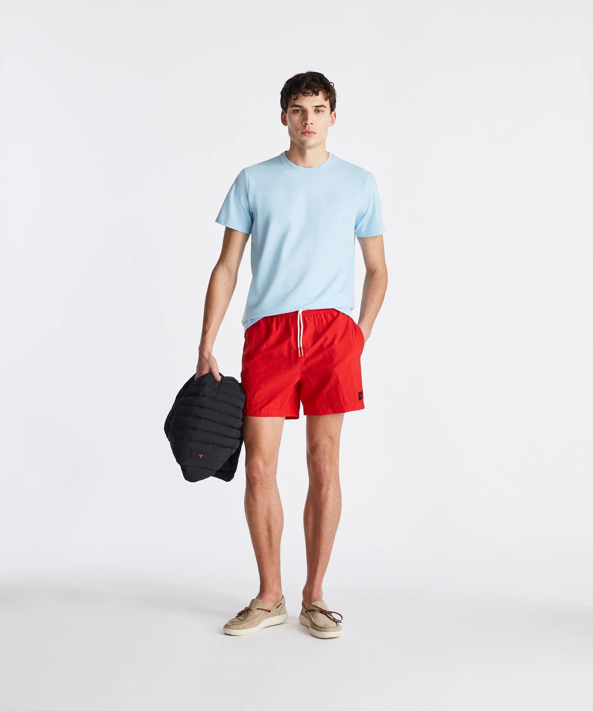 Uomo con t-shirt azzurra, pantaloncini rossi, mocassini beige e giubbino nero in mano