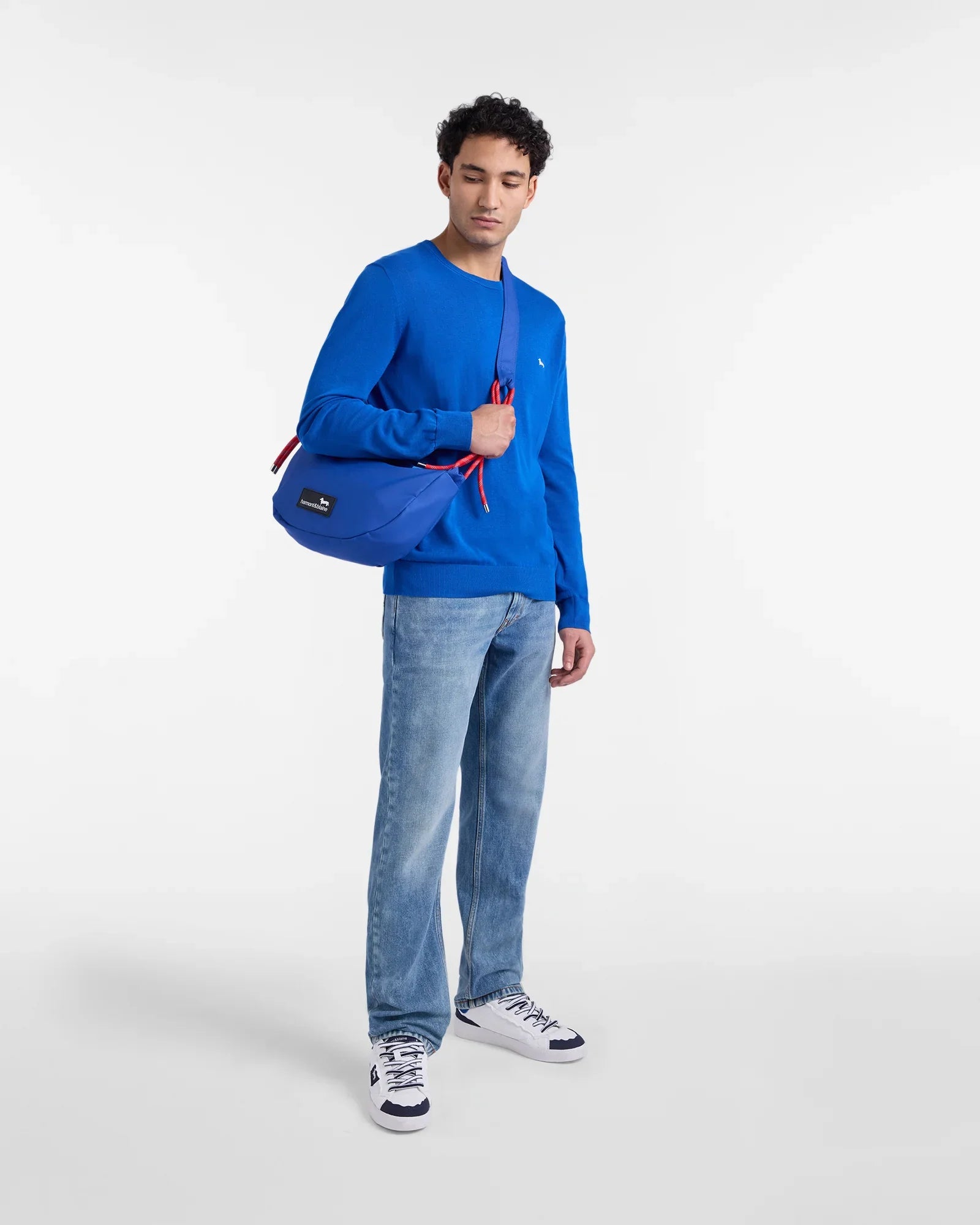 Modello con maglia blu, jeans chiari, sneakers bianche e borsa a tracolla blu, moda uomo.