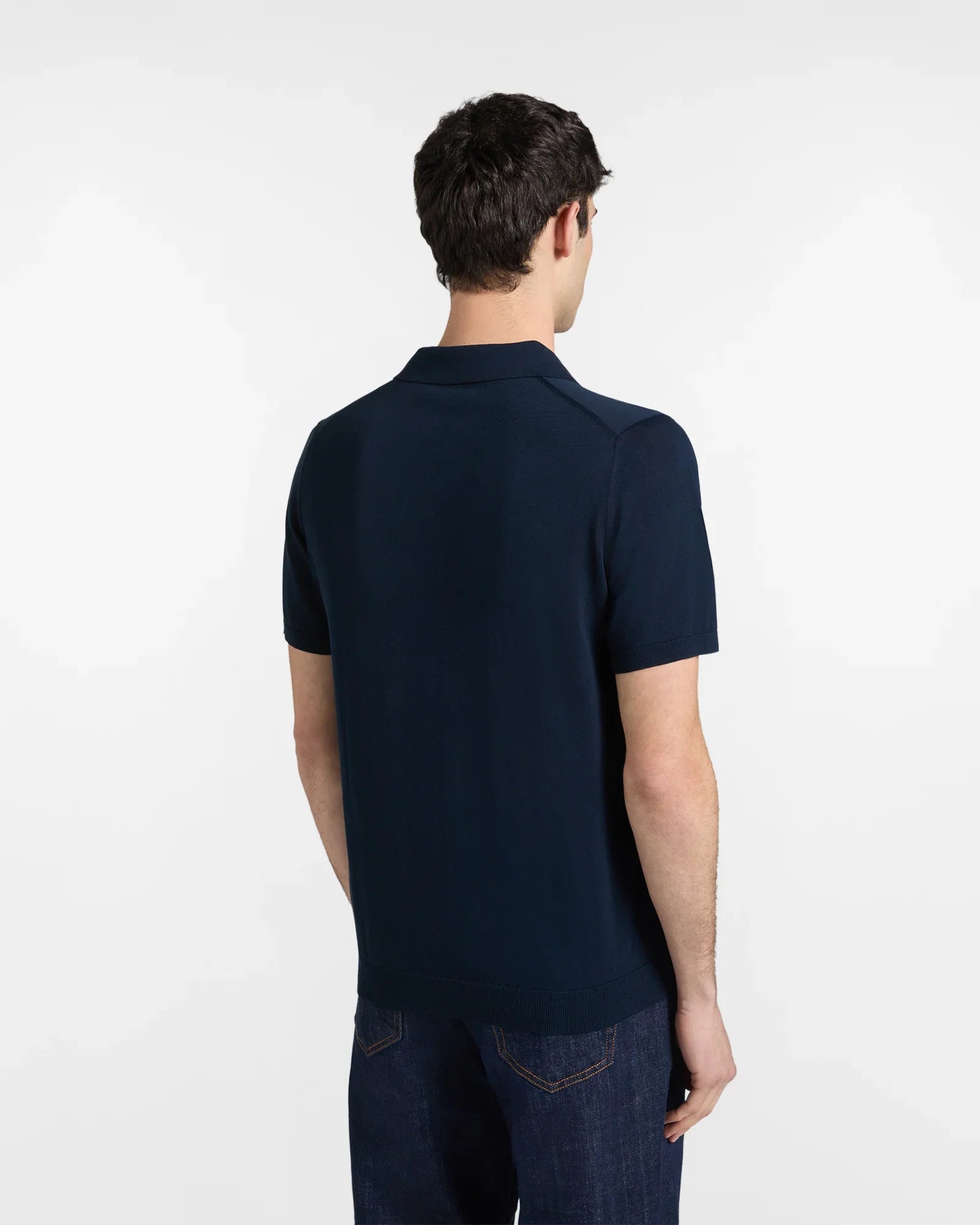 Uomo di spalle con polo blu navy manica corta e jeans, sfondo neutro.