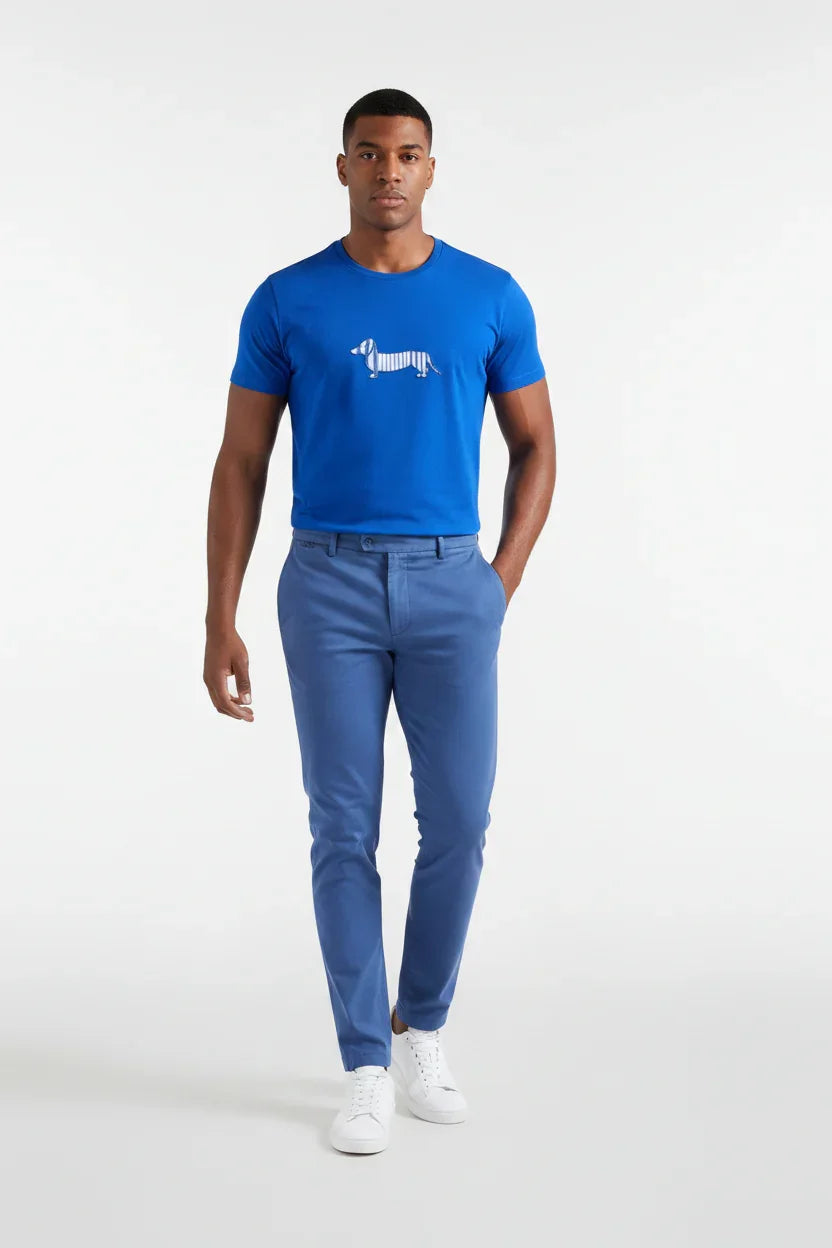 Uomo con t-shirt blu con cane stampato, pantaloni blu e sneakers bianche su sfondo neutro