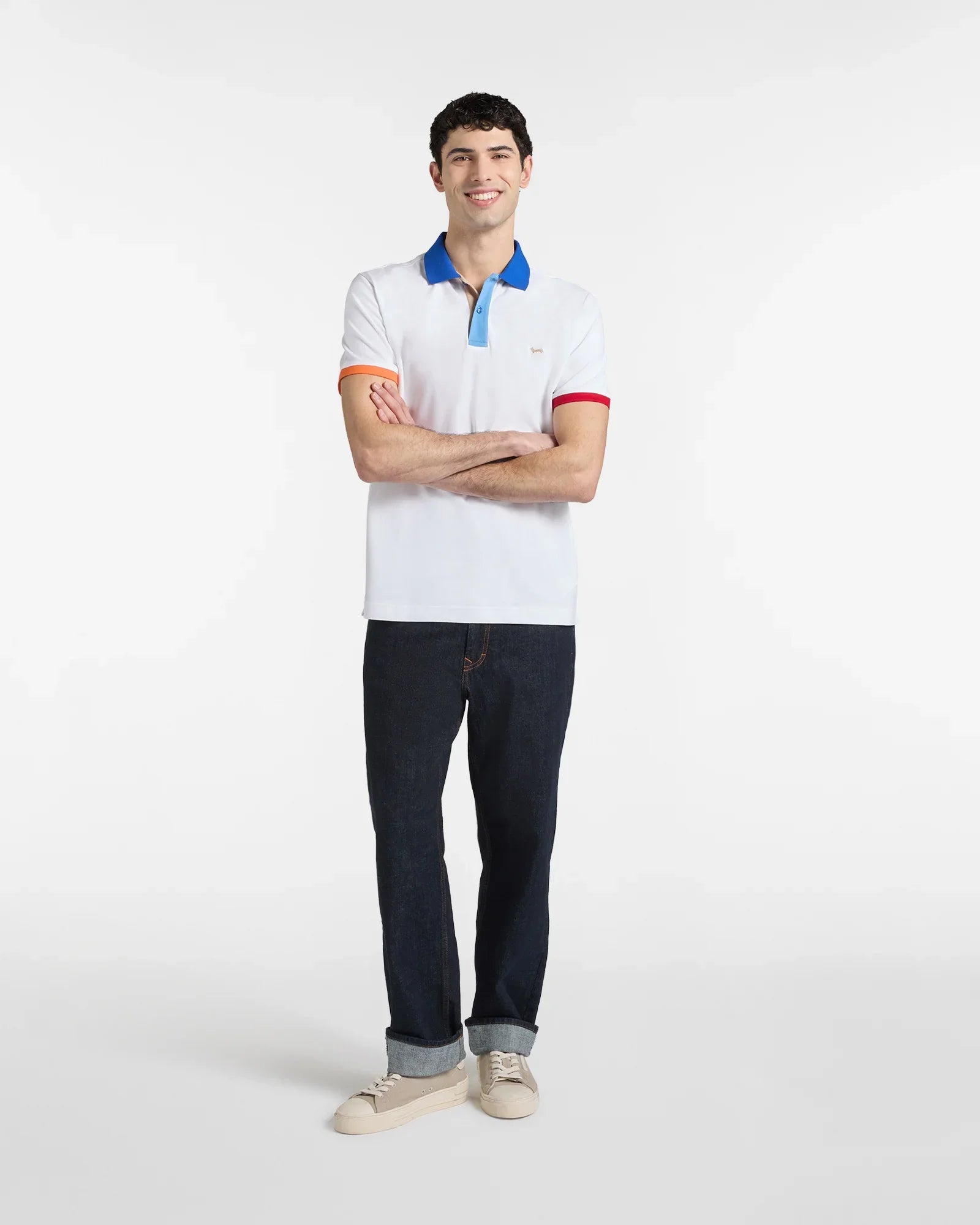 Modello uomo con polo bianca dettagli colorati, jeans scuri e sneakers beige su sfondo neutro