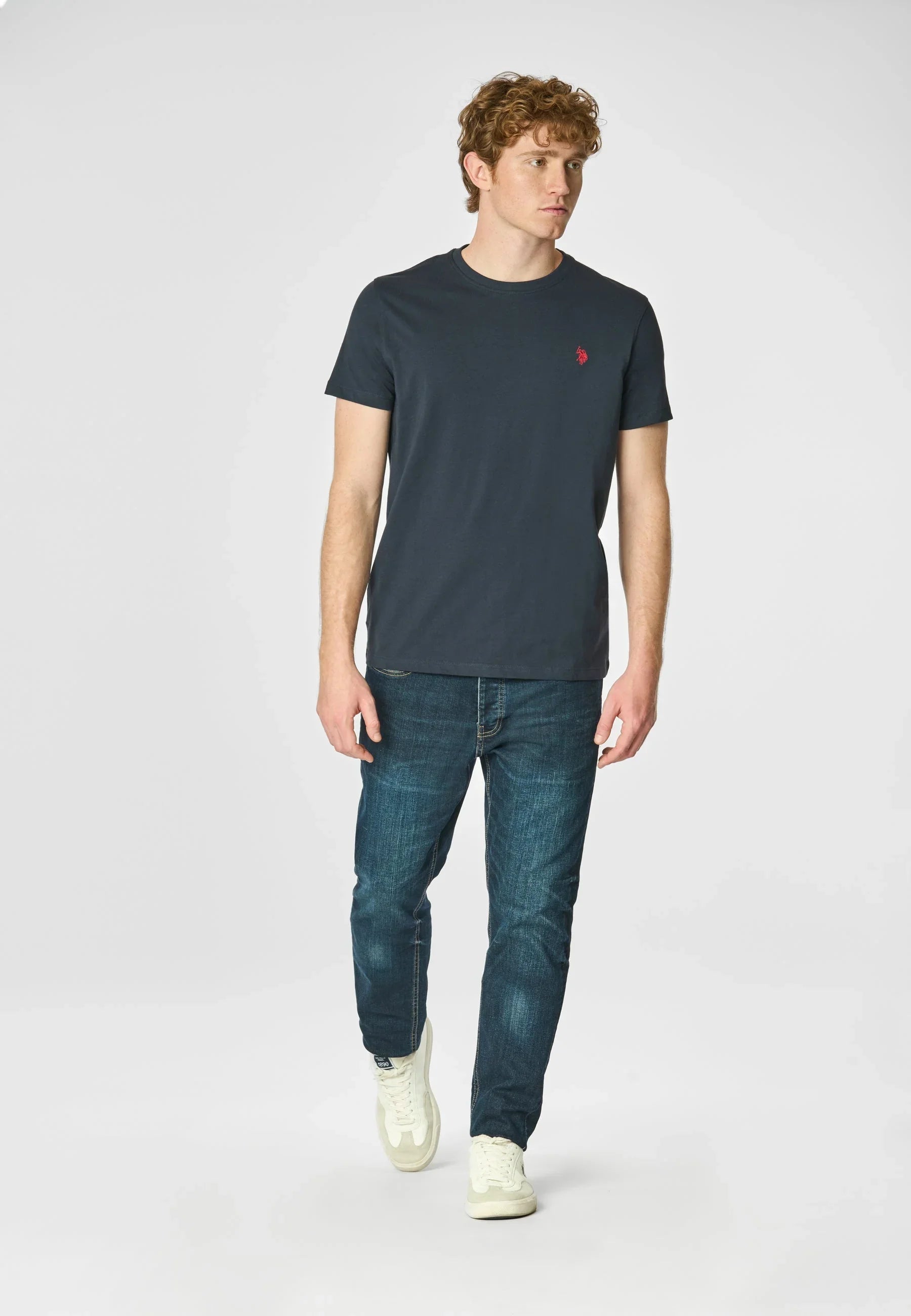 Modello con t-shirt blu scuro con logo rosso, jeans denim e sneakers bianche uomo.
