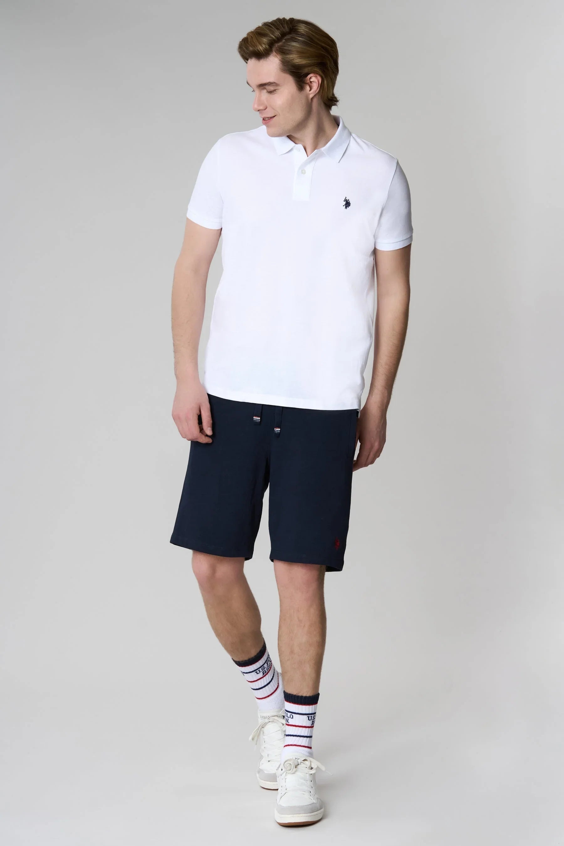 Modello indossa polo bianca e bermuda blu uomo, stile casual estivo, sfondo neutro.