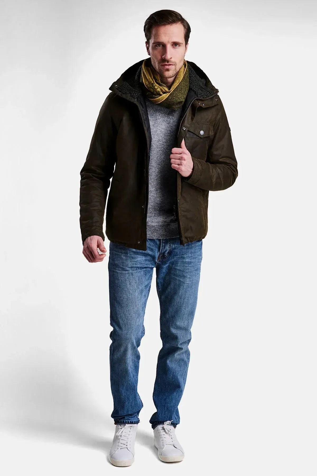 Giacca cerata verde uomo, sciarpa gialla, jeans blu, sneakers bianche, look casual elegante.