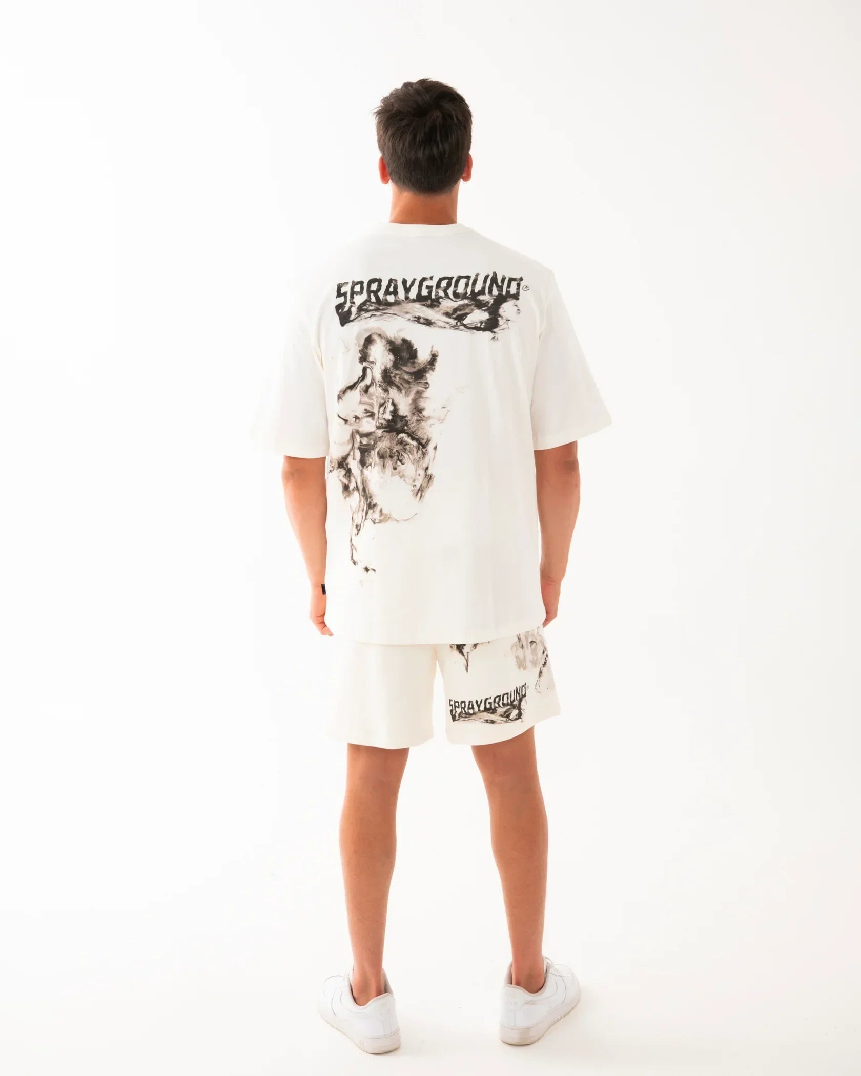 Outfit uomo estivo con t-shirt e bermuda bianchi Sprayground, stampa nera, look casual moderno.