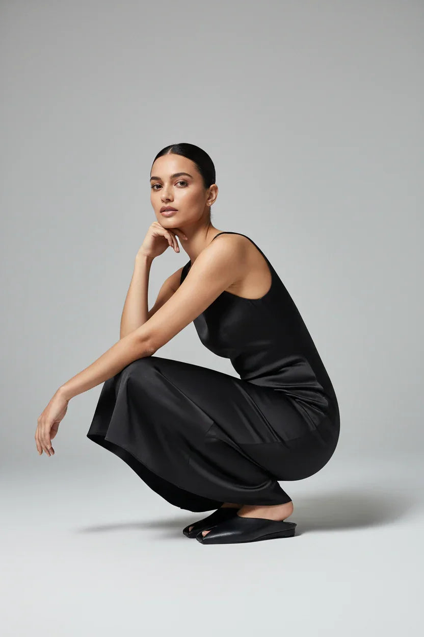 Donna elegante in abito nero satinato e sandali minimal, posa su sfondo neutro.